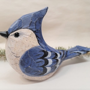 May include: Hand-carved wooden bluebird figurine with white and black accents. The bird has a blue crest and a white belly.