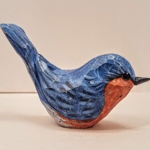May include: Hand-carved wooden bluebird figurine with a red breast and black eyes. The bird is perched on a white surface.