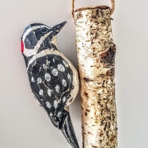 May include: A hand-painted wooden bird ornament with black and white markings and a red beak, hanging from a birch branch.