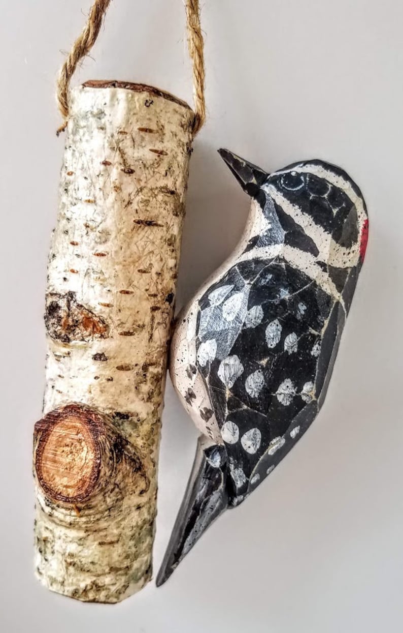 May include: A wooden bird ornament, painted black and white with white spots, is hanging from a piece of twine. The bird is perched on a small, wooden birch log ornament, also hanging from twine.