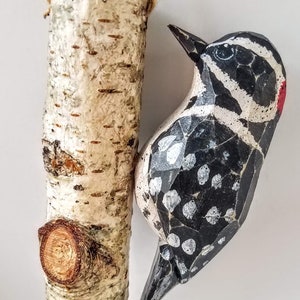 May include: A wooden bird carving of a black and white woodpecker with red accents, hanging from a birch bark branch with a twine loop.