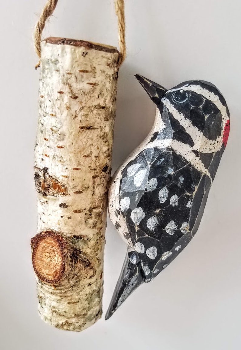 May include: A wooden bird carving of a black and white woodpecker with red accents, hanging from a birch bark branch with a twine loop.