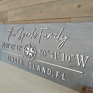 May include: A gray wooden sign with white lettering. The sign reads "The Sparks Family" above coordinates and "SINGER ISLAND, FL". A compass rose is centered on the sign. The sign is displayed against a wooden background.