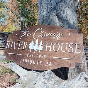 May include: A wooden sign with the words "The Oliver's River House" in white lettering. The sign also includes the text "EST. 2020" and "Tidioute, PA". The sign is brown and has a decorative shape with a tree design.