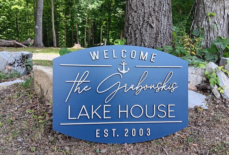 May include: A blue wooden sign with white lettering that reads "Welcome The Grabouskis Lake House Est. 2003". An anchor symbol is above the name.