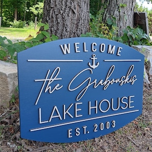 May include: A blue wooden sign with white lettering that reads "Welcome The Grabowski's Lake House Est. 2003". The sign has a white anchor symbol above the name.