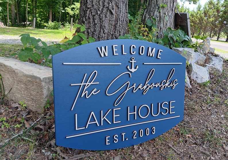 May include: A blue wooden sign with white lettering that reads "Welcome The Grabowski's Lake House Est. 2003". The sign has a white anchor symbol above the name.