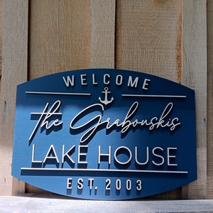 May include: A blue wooden sign with white lettering that reads "Welcome The Grabouskis Lake House Est. 2003". An anchor symbol is above the name.