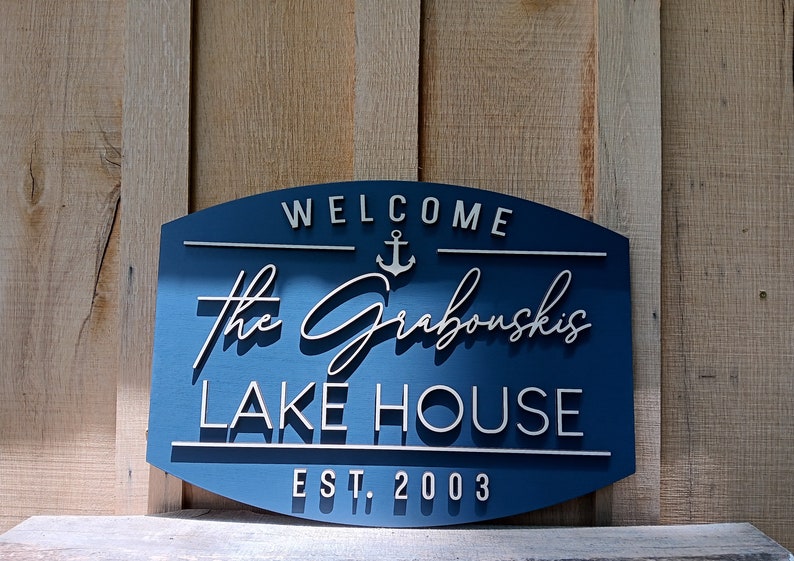 May include: A blue wooden sign with white lettering that reads "Welcome The Grabouskis Lake House Est. 2003". An anchor symbol is above the name.