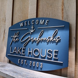 May include: A blue wooden sign with white lettering that reads "Welcome", an anchor symbol, "The Grabowski's Lake House", and "Est. 2003".