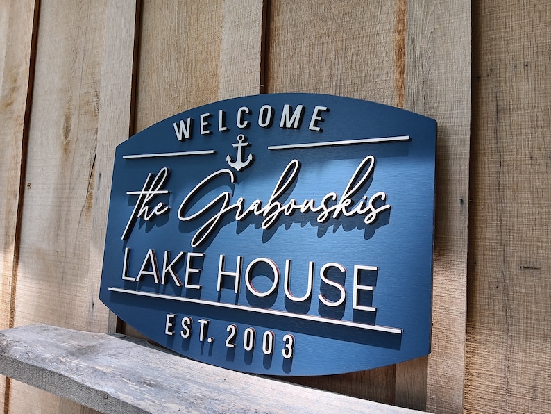 May include: A blue wooden sign with white lettering that reads "Welcome", an anchor symbol, "The Grabowski's Lake House", and "Est. 2003".