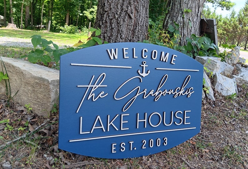 May include: A blue wooden sign with white lettering that reads "Welcome to the Grabouskis Lake House Est. 2003". An anchor symbol is above the name.