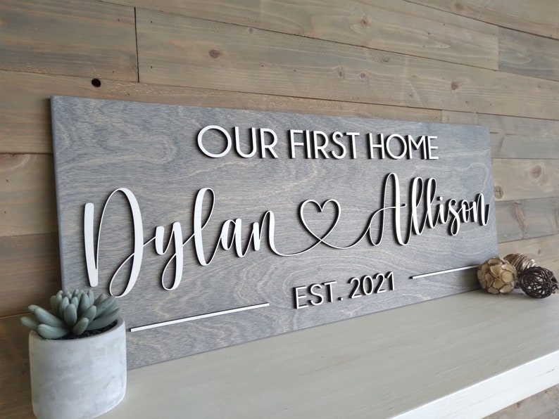 May include: A grey wooden sign with the text "Our First Home Dylan & Allison Est. 2021" in white raised letters.