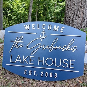May include: A blue wooden sign with white lettering that reads "Welcome The Grabouskis Lake House Est. 2003". An anchor symbol is above the name.