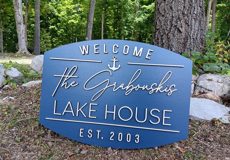 May include: A blue wooden sign with white lettering that reads "Welcome The Grabouskis Lake House Est. 2003". An anchor symbol is above the name.