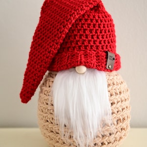 May include: A handmade gnome doll with a red crocheted hat and a long, white faux fur beard. The gnome's body is a beige crocheted sphere. A small wooden bead serves as the nose. The tag reads "CozyCuteCreations".