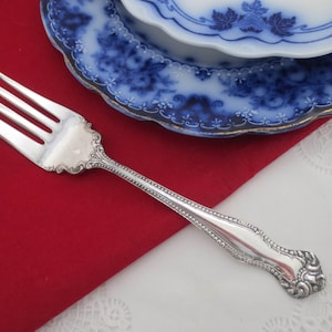 May include: A silver fork with an ornate handle and a tines. The fork is lying on a red surface.