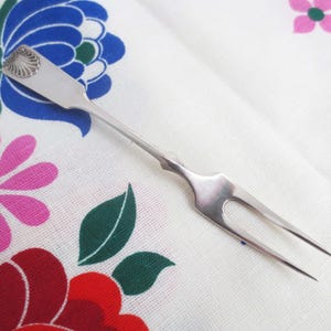 Peut inclure: Une fourchette de service argentée avec un motif de coquillage sur le manche. La fourchette à deux dents est posée sur un fond blanc avec des motifs floraux colorés roses, bleus et rouges.