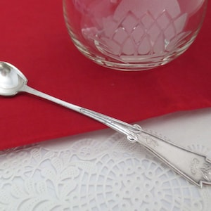 May include: A silver teaspoon with an ornate handle and a rounded bowl. The spoon is lying on a white lace doily.