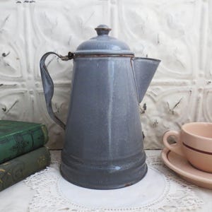 Puede incluir: Una cafetera de esmalte gris con asa y pico. La cafetera tiene un aspecto ligeramente desgastado y está sobre una superficie blanca.