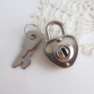 May include: A silver-colored heart-shaped padlock with a matching key. The padlock has a rounded top and a keyhole in the center. The key is flat with a ring at the top. The items are on a white surface.