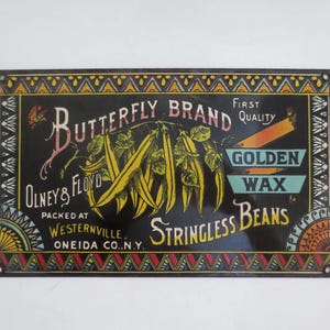 Op de afbeelding: Vintage metalen bord voor Butterfly Brand Golden Wax Stringless Beans. Het bord heeft een zwarte achtergrond met kleurrijke tekst en illustraties van gele bonen. Er staat "Olney & Floyd, Packed at Westernville, Oneida Co., N.Y."