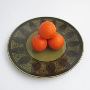 May include: A decorative plate with a green center and a dark green and gold patterned rim. Three bright orange tangerines are arranged on the plate, creating a vibrant contrast. The plate is round and has a vintage aesthetic.