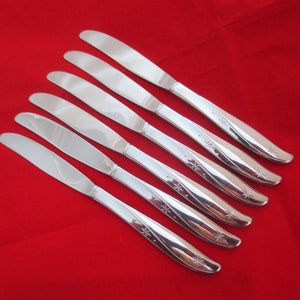 Puede incluir: Seis cuchillos de cena de plata con un diseño de estrella en el mango. Los cuchillos están dispuestos en una fila, con los mangos apuntando hacia la derecha.