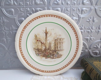 May include: A cream-colored decorative plate featuring a detailed illustration of a London street scene with a tall monument. The plate has a brown and green border and the text "KING WILLIAM STREET, LONDON, 1840".