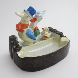 Pode incluir: Um cinzeiro de cerâmica vintage com uma família de patos. Os patos são pintados com cores vivas e usam chapéus e cachecóis. O cinzeiro é castanho com interior branco e tem vários suportes para cigarros.