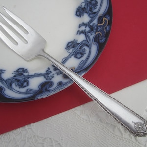 May include: A silver fork with four tines rests on a white plate with a blue floral design. The plate sits on a red surface, with a white lace-like fabric in the background. The fork's handle has a decorative edge.