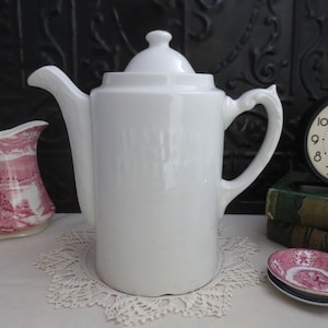 May include: A white ceramic coffee pot with a lid, spout, and handle. The pot is on a white lace doily. A small pink and white jug and a small plate are also visible.