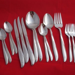 May include: A collection of polished stainless steel silverware, including spoons, knives, and forks, arranged on a red surface. The silverware features a starburst design at the base of the handles. The set includes various sizes of spoons, knives, and forks.