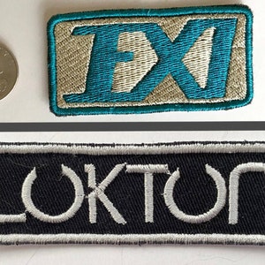 Puede incluir: Dos parches bordados. El parche superior tiene las letras "FX" en color turquesa sobre un fondo beige. El parche inferior dice "LOKTON" en blanco sobre un fondo negro. Una moneda de 2,5 cm está al lado del parche superior para comparar el tamaño.