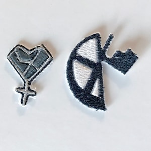 Op de afbeelding: Twee geborduurde patches. Eén is een grijs hart met een vuistontwerp en een vrouwelijk symbool. De andere is een zwart-wit vredesteken met een zwart bergontwerp. Beide patches hebben witte randen.