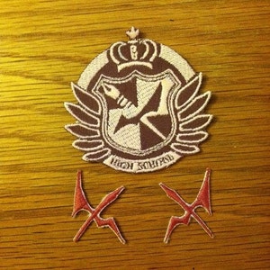 Könnte beinhalten: Zwei rote gestickte Patches in Form eines X und ein braunes und weißes gesticktes Patch mit einer Krone, Flügeln und einem Schild mit einer Feder und einem Buchstaben X. Der Schild trägt den Text "Hope's Peak".