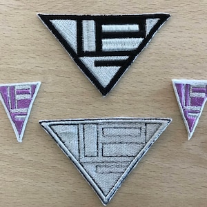 Op de afbeelding: Drie geborduurde strijkpatches in de vorm van driehoeken. De bovenste patch is zwart-wit met een geometrisch ontwerp. De twee onderste patches zijn paars en wit met een vergelijkbaar geometrisch ontwerp.