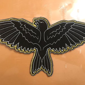 May include: Black embroidered patch featuring a stylized bird with outstretched wings. The bird is outlined in white and yellow stitching, with detailed wing patterns. The patch is designed to be ironed on or sewn onto fabric, suitable for jackets, bags, or other apparel.