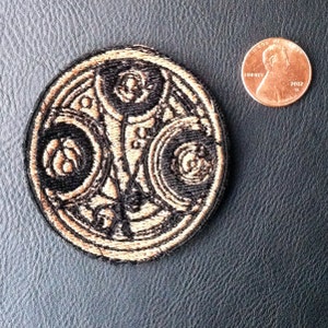 Pode incluir: Um remendo bordado preto e dourado com um design circular com um símbolo no centro. O símbolo é cercado por círculos concêntricos.