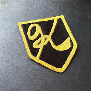 May include: A black and yellow embroidered patch in the shape of a shield with the letters "GF" in a stylized script.