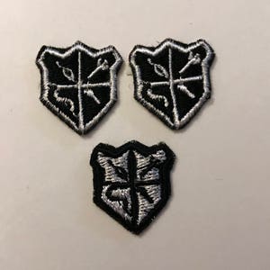 May include: Three black and white embroidered iron-on patches in the shape of a shield. Each shield has a different design with a cross in the center.