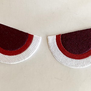 May include: Two embroidered patches in a half-circle shape, resembling eyes. Each patch features a white outer edge, a red middle band, and a dark burgundy center. The patches are designed to be sewn onto fabric.