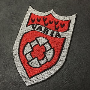 Może przedstawiać: Naszywka haftowana w kolorze czerwonym i srebrnym z napisem "VANIA" czarnymi literami i stylizowanym wzorem koła z ośmioma punktami.