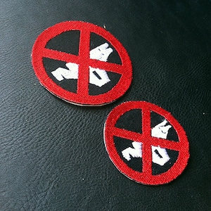 May include: Two embroidered patches with a red border and a black center. A red cross divides the center, with white shapes and the word "NO" in white. The patches are circular and sit on a black textured surface.
