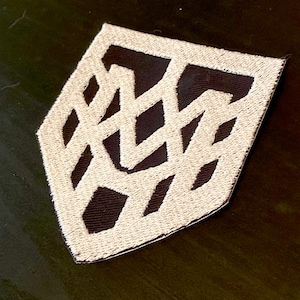 Puede incluir: Parche bordado con un diseño geométrico. El parche presenta un patrón crema entrelazado sobre un fondo marrón oscuro. El parche tiene forma de diamante con una parte inferior puntiaguda.