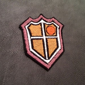 以下が含まれることがあります： 白い縁取りのある刺繍シールドパッチ。十字のデザインとオレンジ色の円が金色の背景に配置されています。パッチはピンクと黒の縁取りがあり、衣類やアクセサリーに適しています。