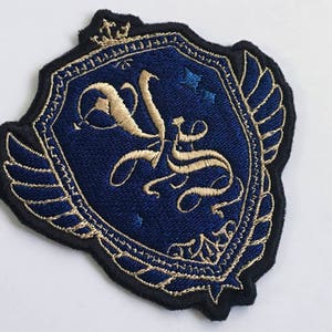 Puede incluir: Parche bordado con un diseño de escudo. El parche presenta un fondo azul marino con letras y detalles dorados. Un borde negro rodea el escudo y una pequeña corona está en la parte superior. El parche tiene una estética vintage o heráldica.