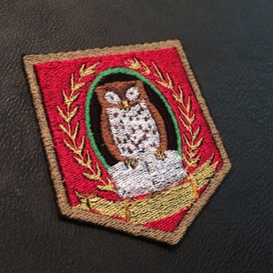May include: Embroidered patch featuring a brown owl within a green wreath on a red background. The owl is surrounded by gold leaves and a gold banner.