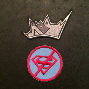 以下が含まれることがあります： 2つの刺繍パッチ。1つ目は、グレーの家が内側に描かれたシルバーのクラウン。2つ目は、赤い円の中に青いスーパーマンのシンボルが描かれており、赤い線で消されています。