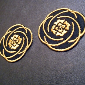 May include: Two black fabric patches with gold embroidered floral designs. The patches are circular and feature a stylized flower with multiple layers of petals.